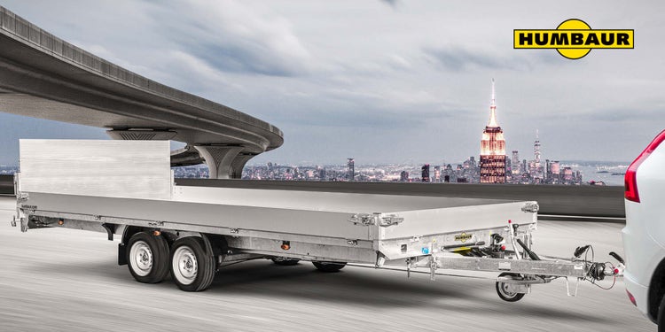 Humbaur Fahrzeuganhänger auf Autobahn mit Stadtpanorama im Hintergrund.