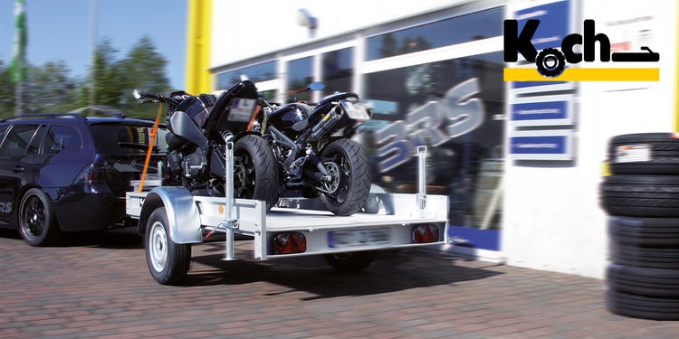 Ein Koch Motorradanhänger steht vor einer Werkstatt mit 2 Motorrädern drauf