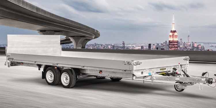 Humbaur Fahrzeuganhänger auf Autobahn mit Stadtpanorama im Hintergrund.